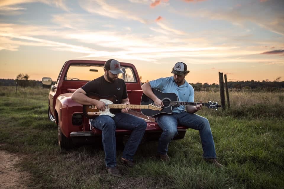 Will and Reid Shackelford playing guitars on a red pickup tailgate at sunset